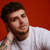 A man in a white tshirt with short brown hair in front of a red background